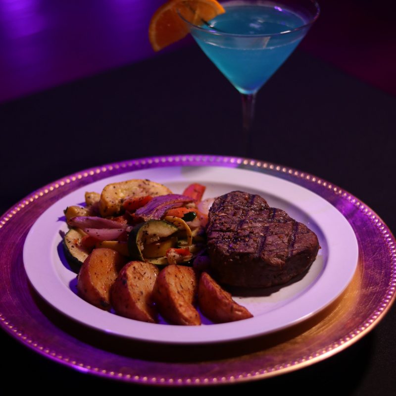 Steak with potatoes and vegetables on a plate, cocktail beside it.