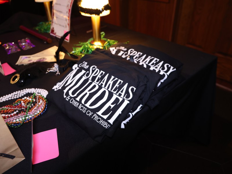 Table with 'A Speakeasy Murder' T-shirts and party items.