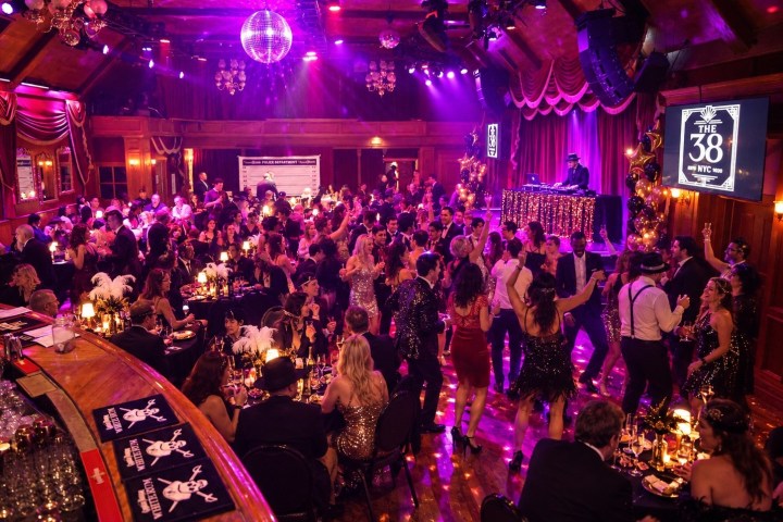 Crowded dance floor with people in formal attire, colorful lighting, disco ball, and a DJ booth on stage.