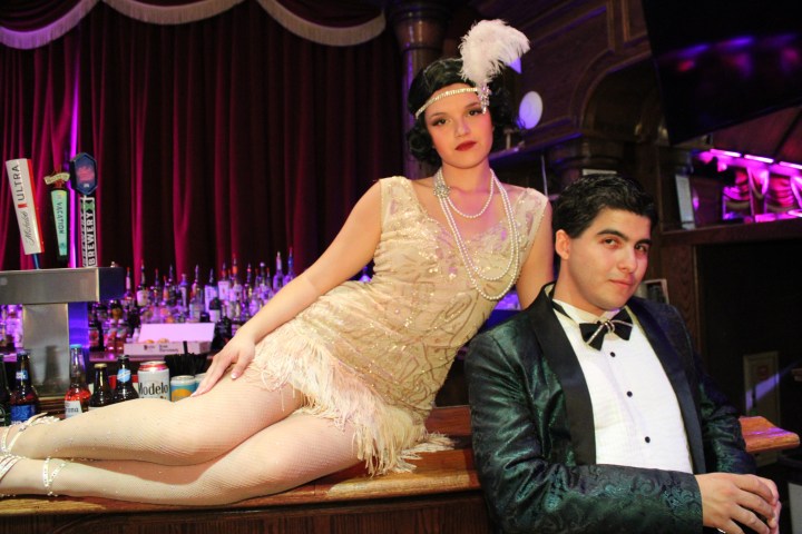 Two people in 1920s attire at a bar; woman in flapper dress, man in tuxedo.