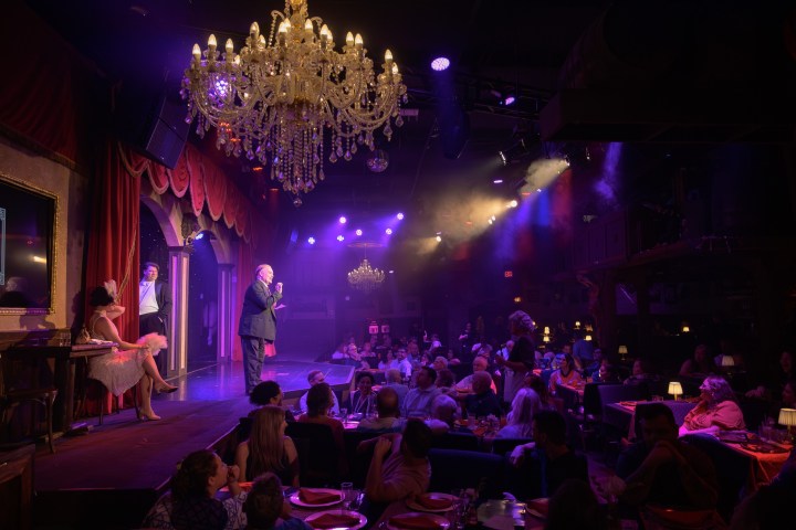 Performer on stage in a dimly lit theater with a chandelier.