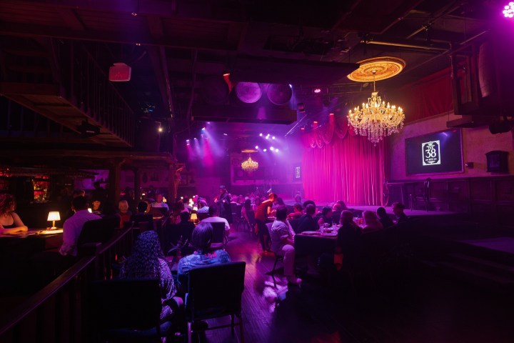 Dimly lit venue with tables, stage, chandeliers, and purple lighting.