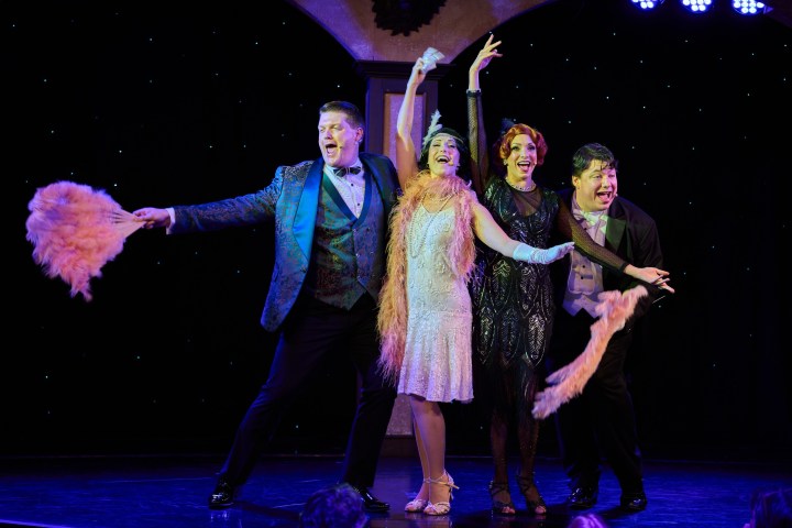 Four performers in vintage costumes singing and dancing with feather props on stage.